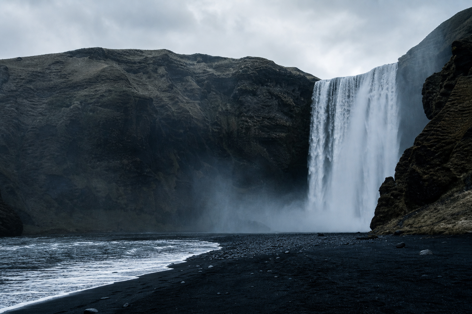 Waterfall cascading beside a black-sand beach in Iceland with soft misty light