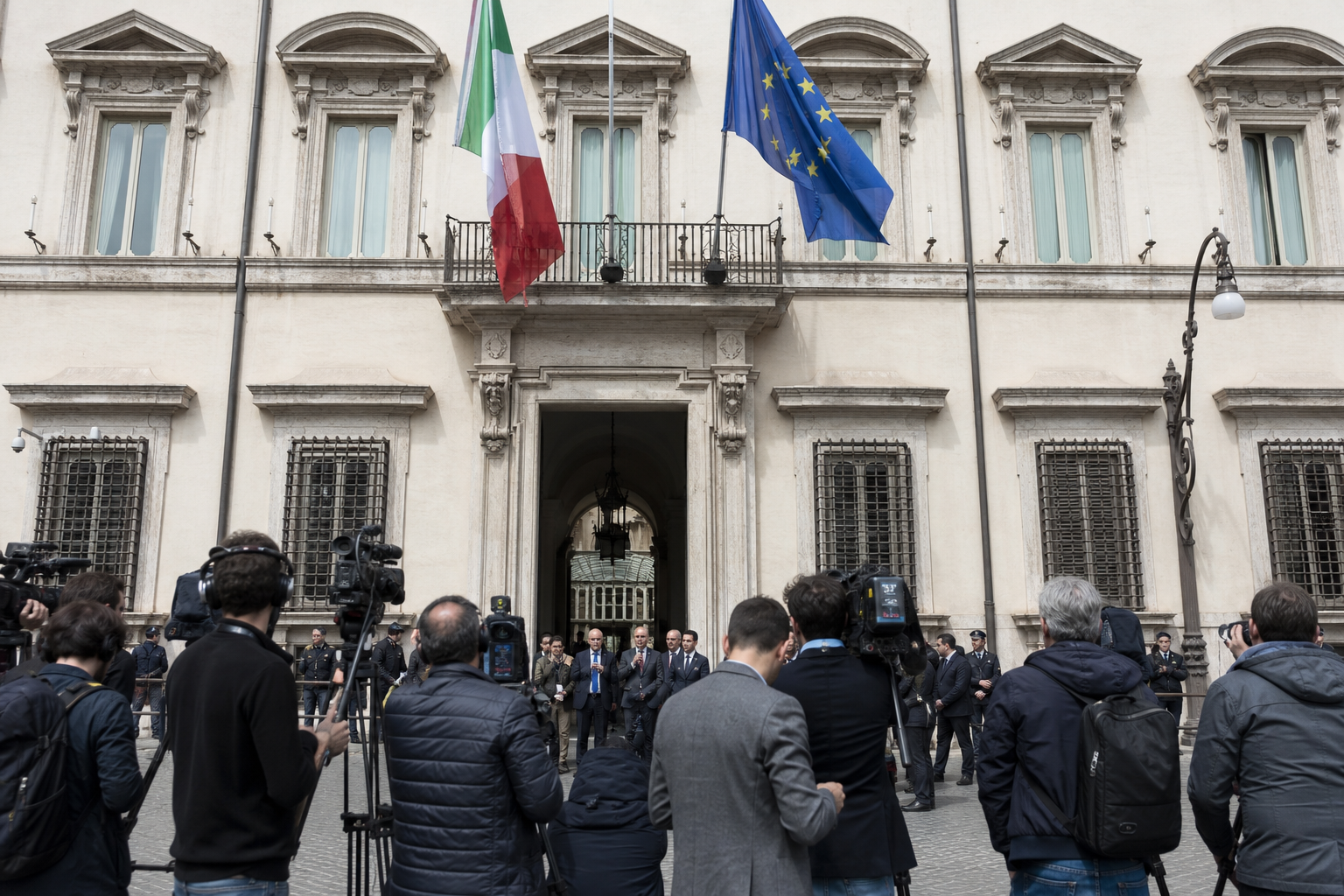 Facciata istituzionale con bandiere e giornalisti davanti all'ingresso durante una conferenza politica